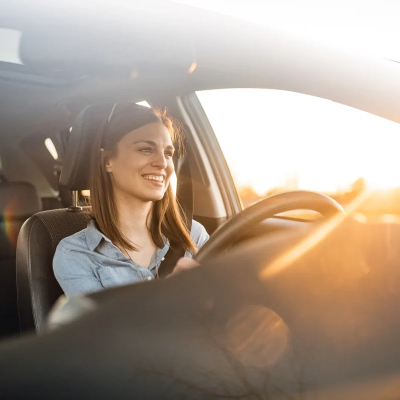 A smiling woman in a denim shirt drives a car with one hand on the wheel as sunset light streams through the windows.