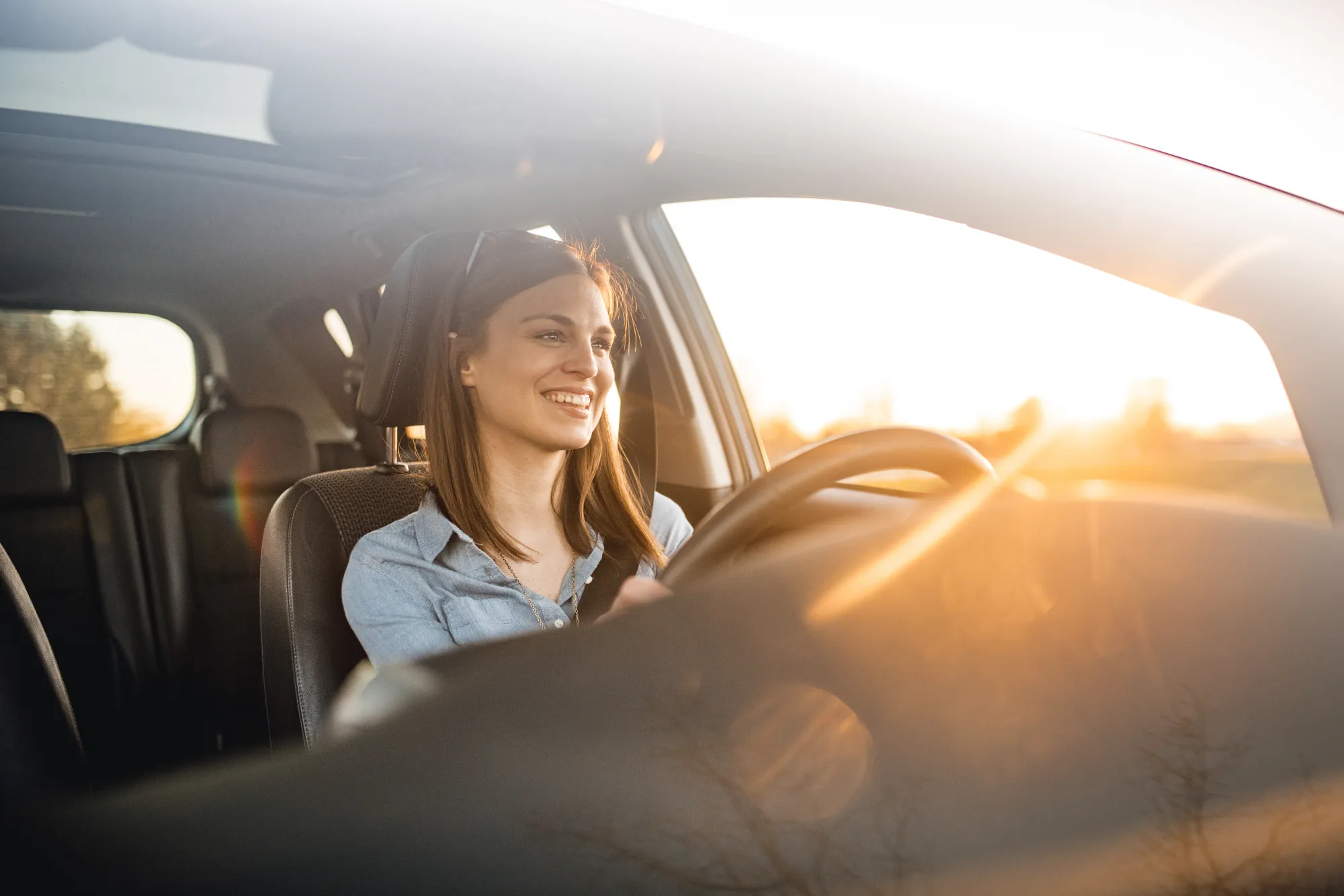 A smiling woman in a denim shirt drives a car with one hand on the wheel as sunset light streams through the windows.