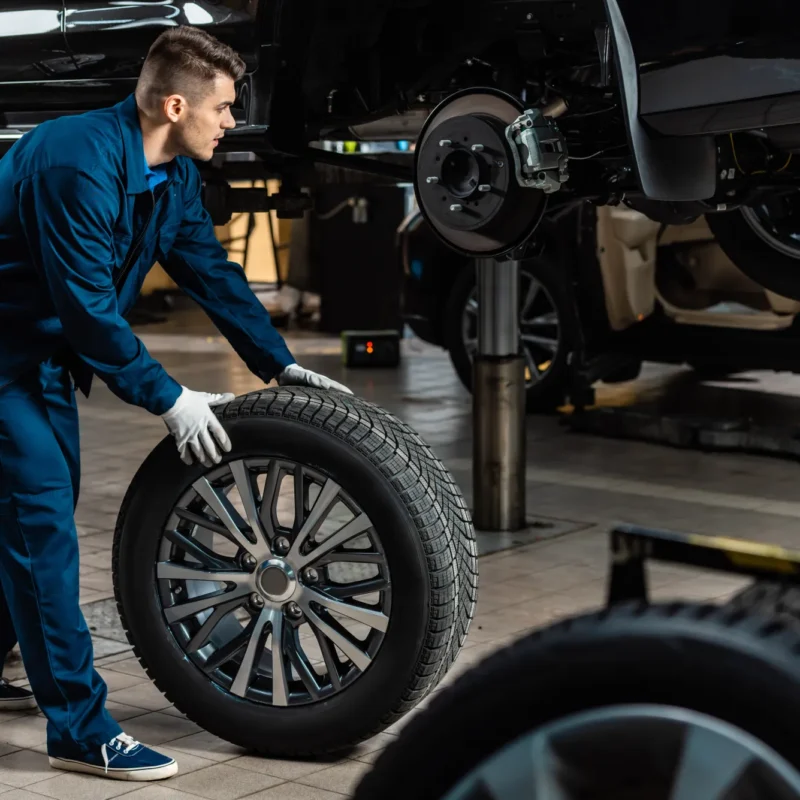 A mechanic in blue coveralls rolls a tire on an alloy wheel toward a vehicle raised on a shop lift with one wheel removed.