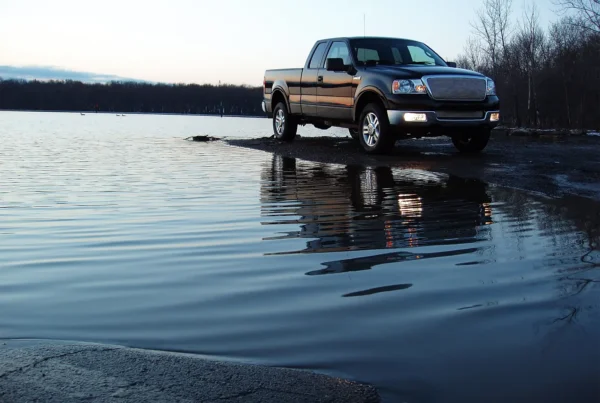 A black pickup truck parked at the edge of a calm lake at dusk, reflected in the water with bare trees along the far shore.