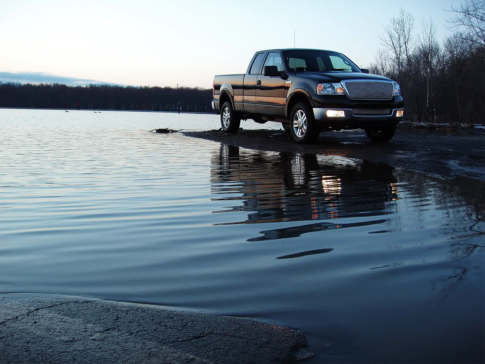 A black pickup truck parked at the edge of a calm lake at dusk, reflected in the water with bare trees along the far shore.