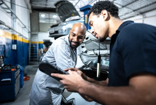 A smiling mechanic and a man in a navy polo look at a tablet together in a repair shop, with a car's open hood behind them.