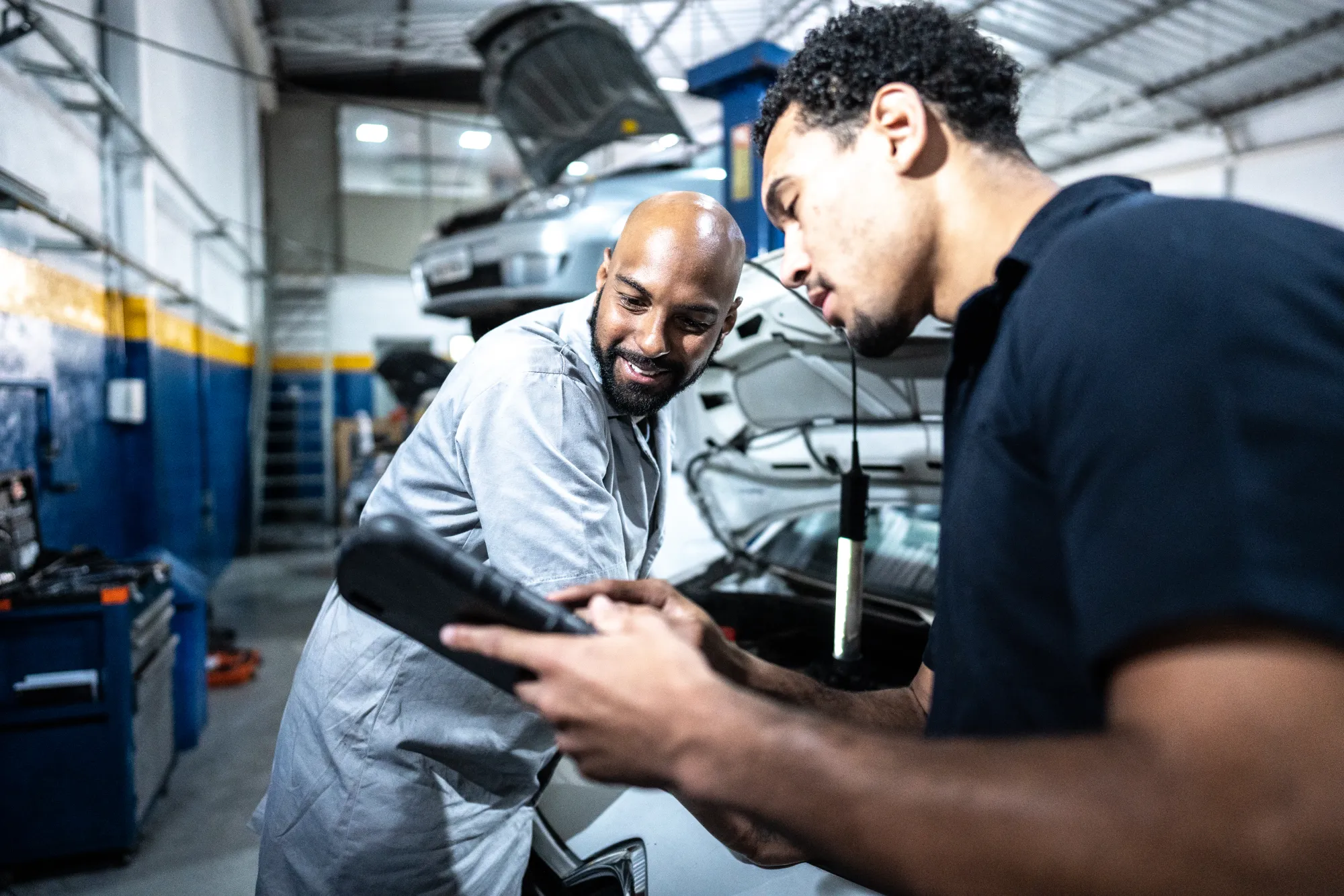 A smiling mechanic and a man in a navy polo look at a tablet together in a repair shop, with a car's open hood behind them.